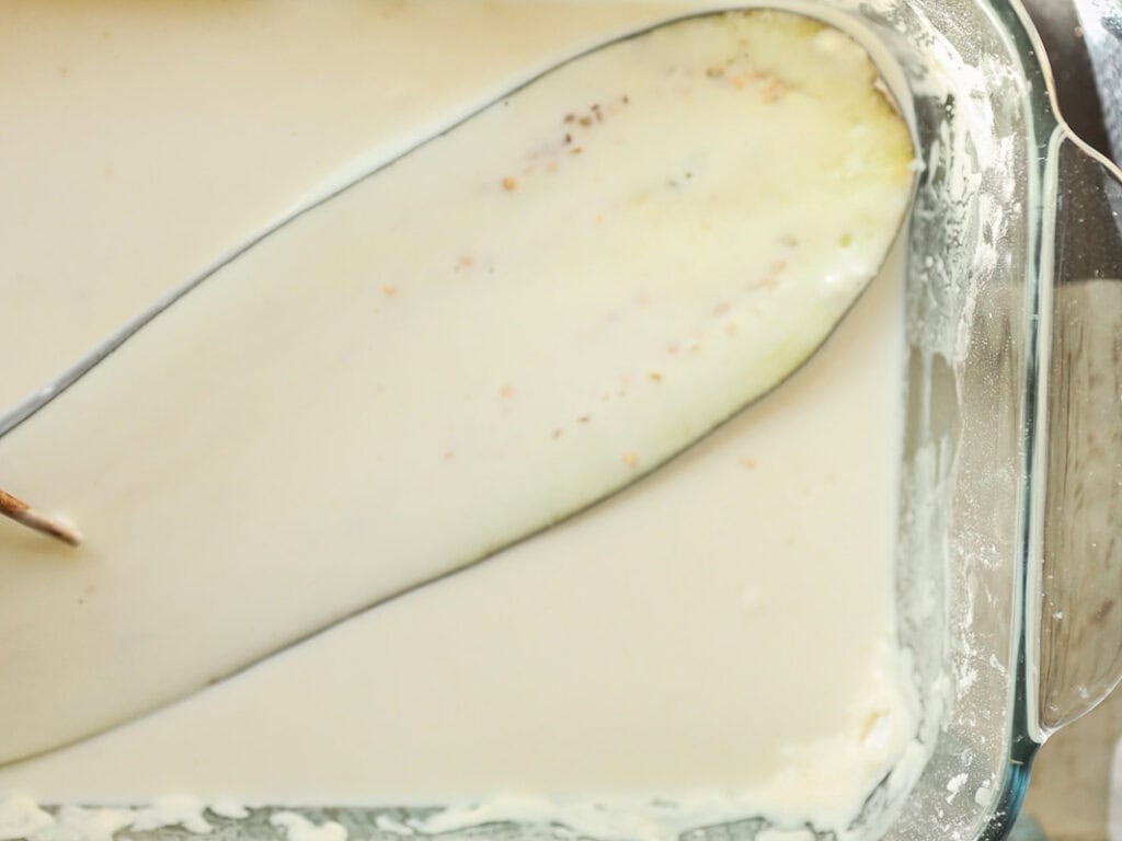 eggplant is dipped into a thick creamy liquid in a glass dish, showing a smooth, pale texture with small specks.